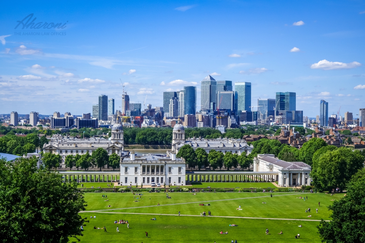 A wide shot of London's skyline from the elevated vantage point of Greenwich Park. The foreground showcases the historic architecture of the Old Royal Naval College with its distinctive twin domes and the elegant Queen's House. The River Thames snakes through the middle ground, leading to the cluster of modern skyscrapers in the City of London, including the recognizable Shard. A bright blue sky dotted with fluffy white clouds completes the scene.