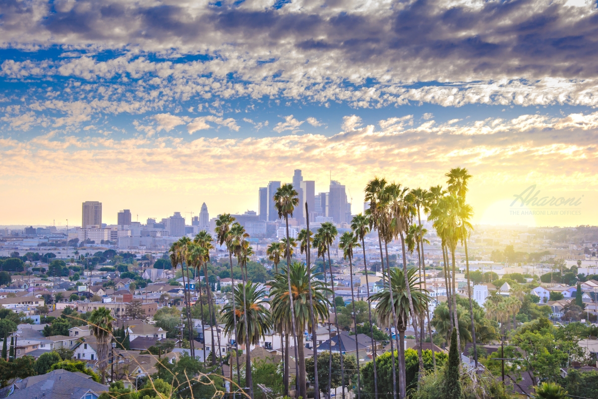 Panoramic view of Los Angeles skyline at sunset, with palm trees framing the cityscape and a vibrant blue sky with scattered clouds above.