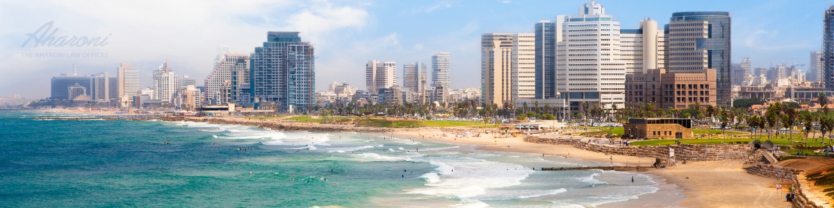 Scenic view of Tel Aviv's beachfront with sunbathers on the beach, waves approaching the shore, and a backdrop of contemporary high-rise buildings against a blue sky.