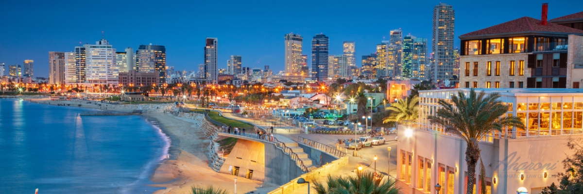 A nighttime panoramic view of Tel Aviv, Israel. The skyline is illuminated with a variety of lights, showcasing modern high-rise buildings. A brightly lit promenade stretches along the coastline, where people can be seen walking. Palm trees dot the landscape, adding to the vibrant urban scene. The calm waters of the Mediterranean Sea reflect the city lights.